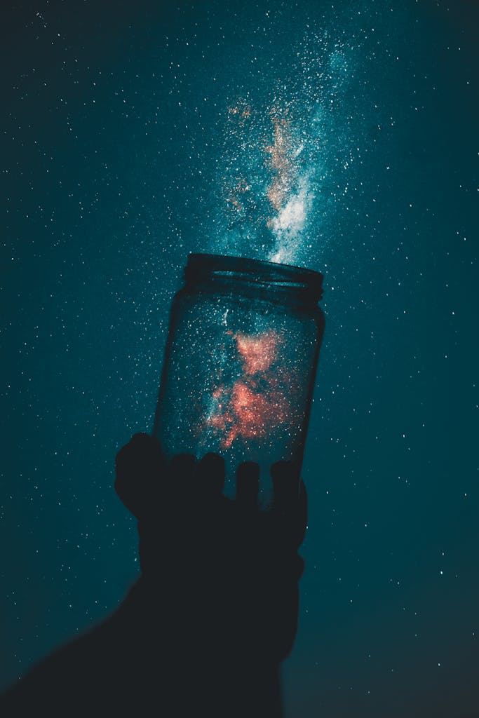 A silhouette hand holds a mason jar capturing a stunning galaxy under a starry night sky.